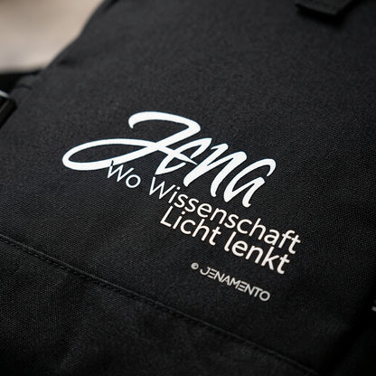 Detailaufnahme des Rolltop Rucksacks Jena.Lichtschrift mit weißem Jena Design auf schwarzem Material. Nahaufnahme von Druck, Stoffstruktur und Verarbeitung.